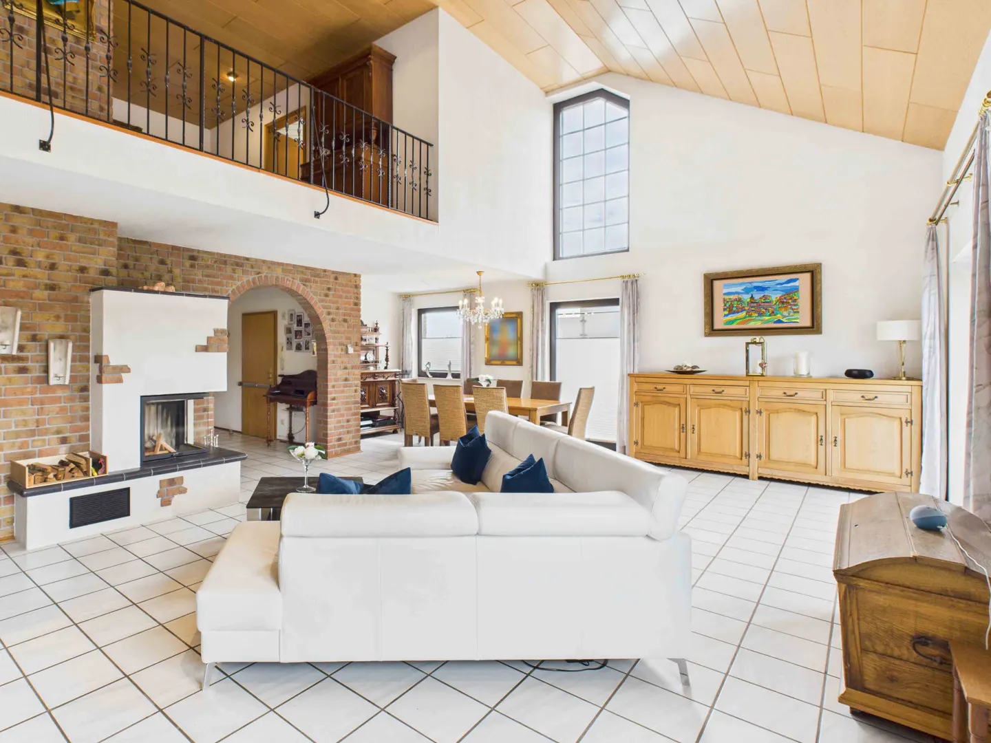 Bright, open living room with a white sofa, brick fireplace, and dining area. A wooden ceiling and large window add natural light.