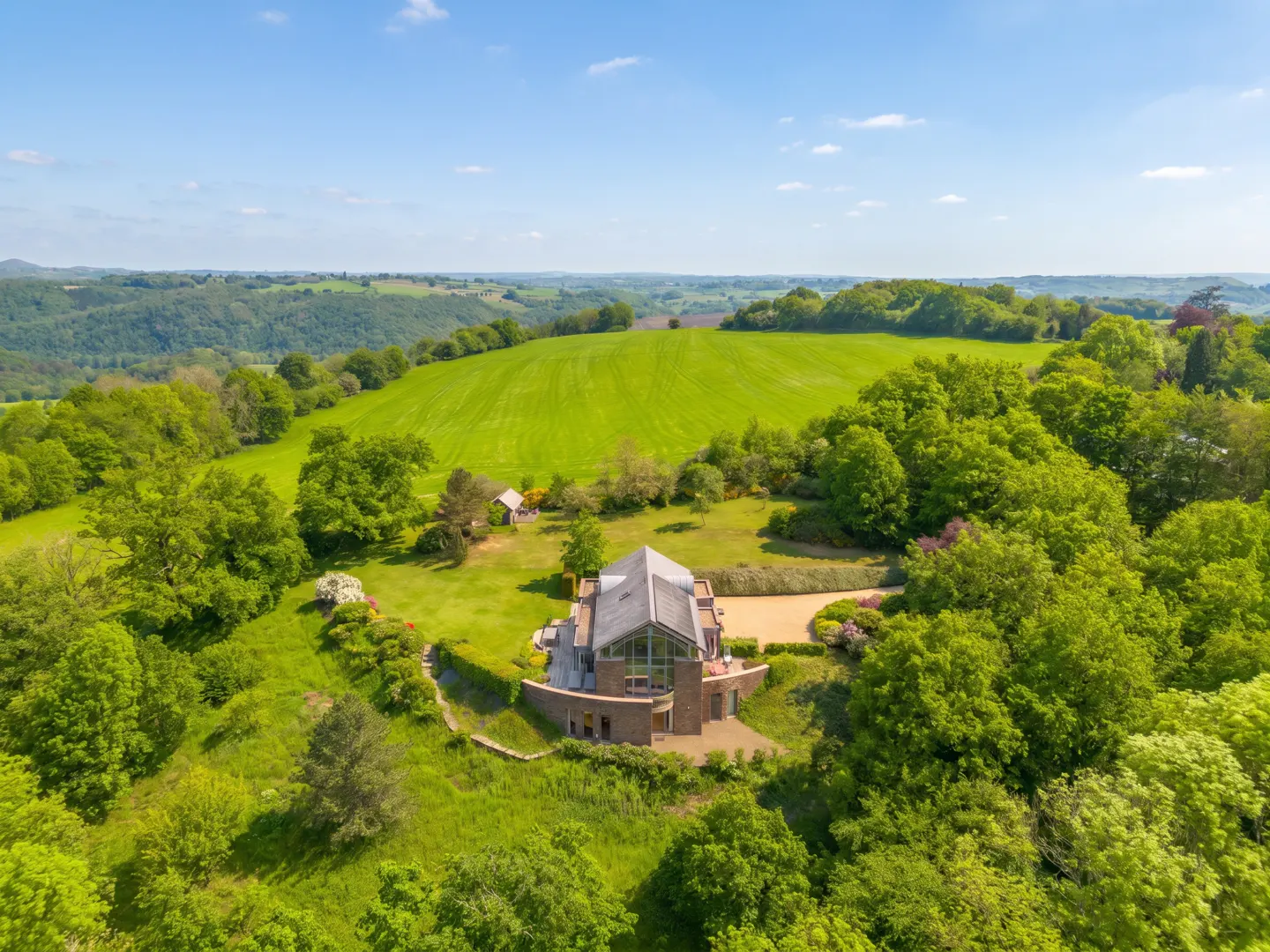 Aerial view of a modern stone house with a glass roof, surrounded by green trees and a large green field under a blue sky.
