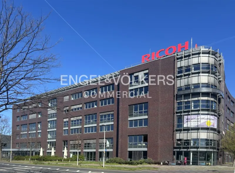 Exterior of a brick office building with many windows under a blue sky. A red "RICOH" sign is on the roof. "Engel & Völkers Commercial" is overlaid.