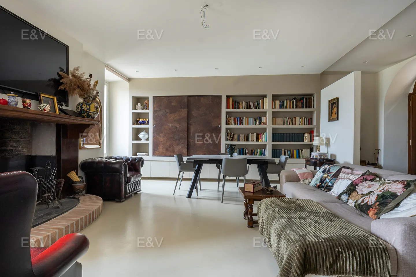 Living room with fireplace, bookshelves, dining table, and sofa. The walls are light beige and the floor is white.
