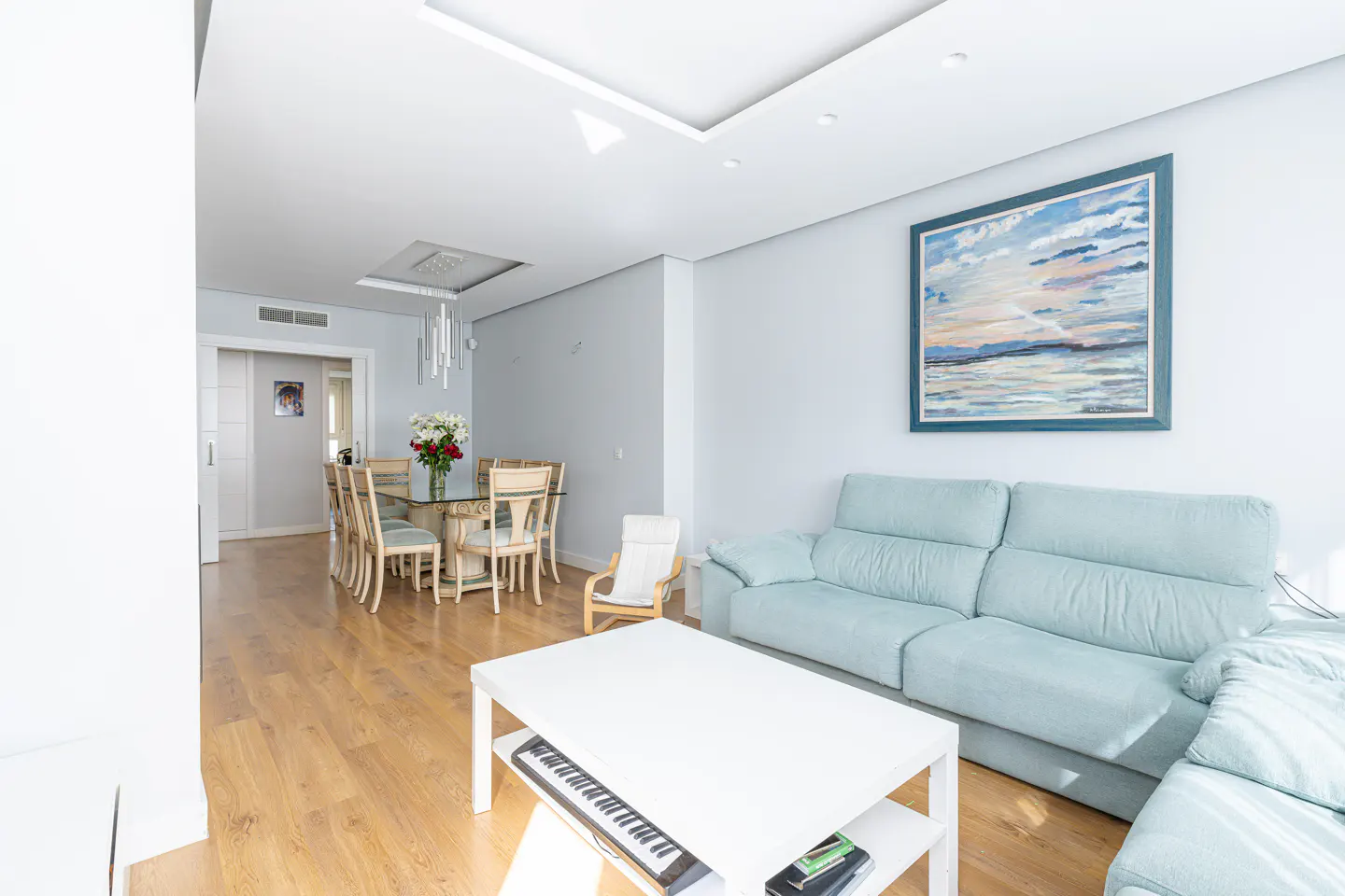 Bright living and dining area with light blue sofa, white coffee table with keyboard, and glass dining table with beige chairs.