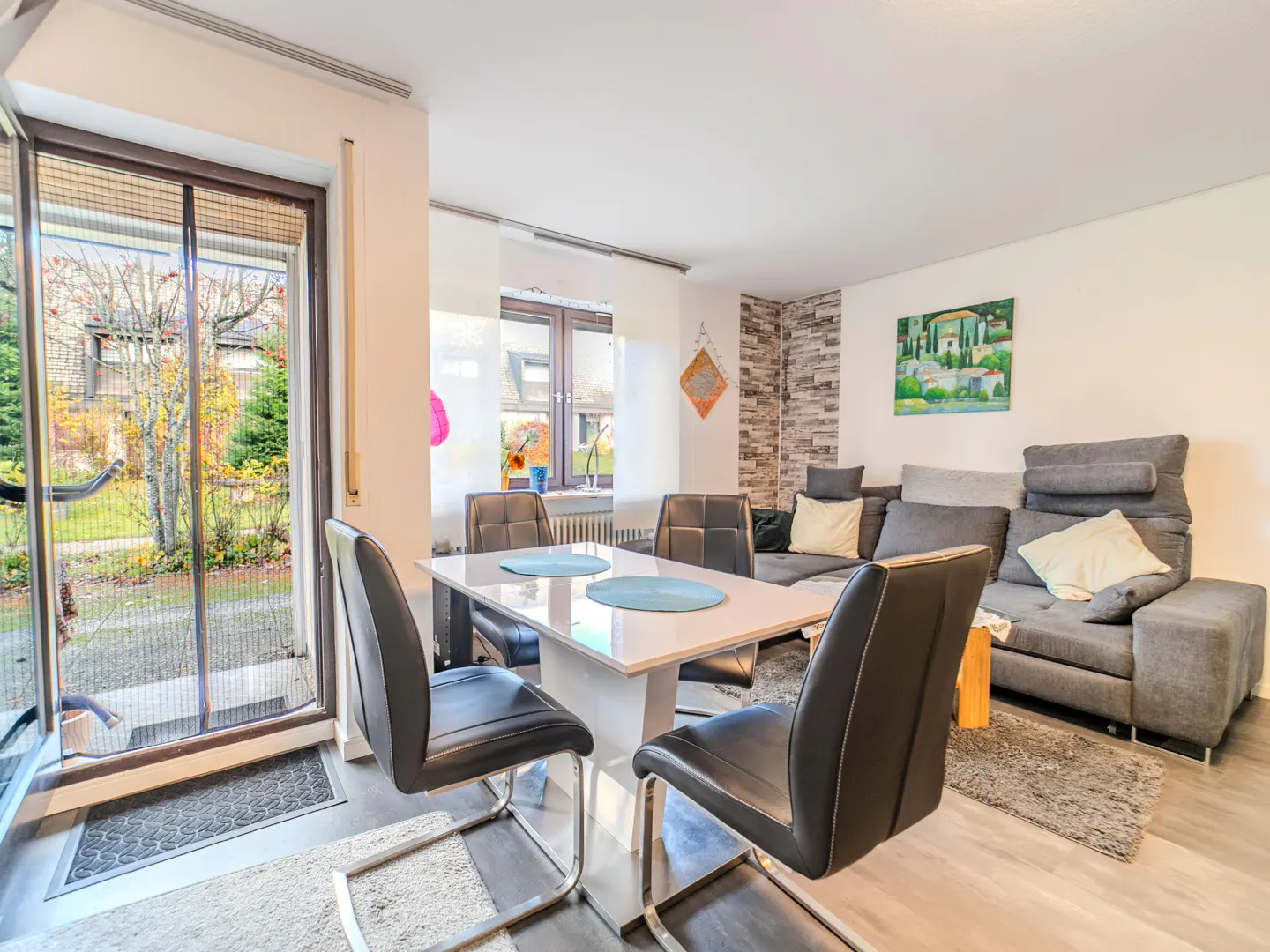 Bright living room with a white table, black chairs, and a gray sofa. A glass door leads to a garden.