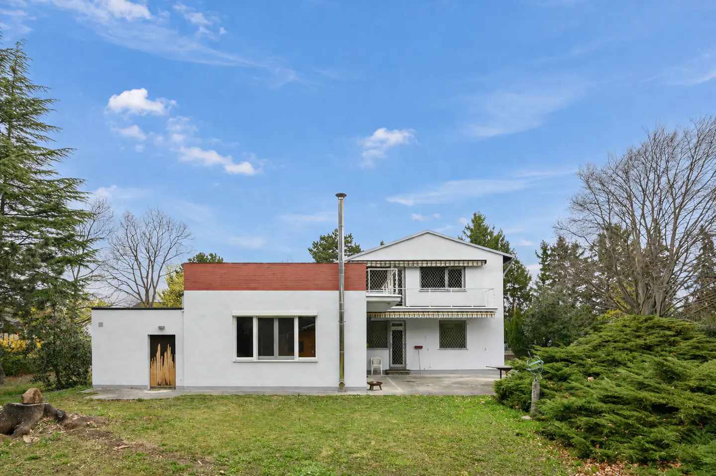Two-story white house with a red brick accent wall, a metal chimney, and a balcony, set against a blue sky with scattered clouds.