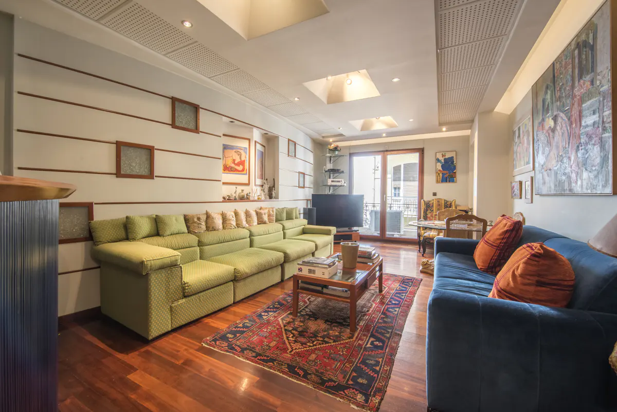 Living room with hardwood floors, a green sectional sofa, a blue sofa with orange pillows, and a patterned rug. Artwork adorns the walls.