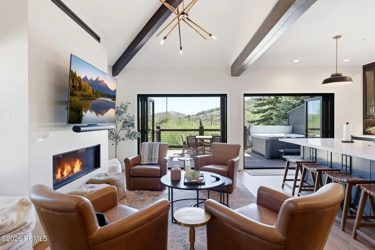 Bright living room with four leather chairs, fireplace, TV, and open doors to a deck with a hot tub and mountain view.