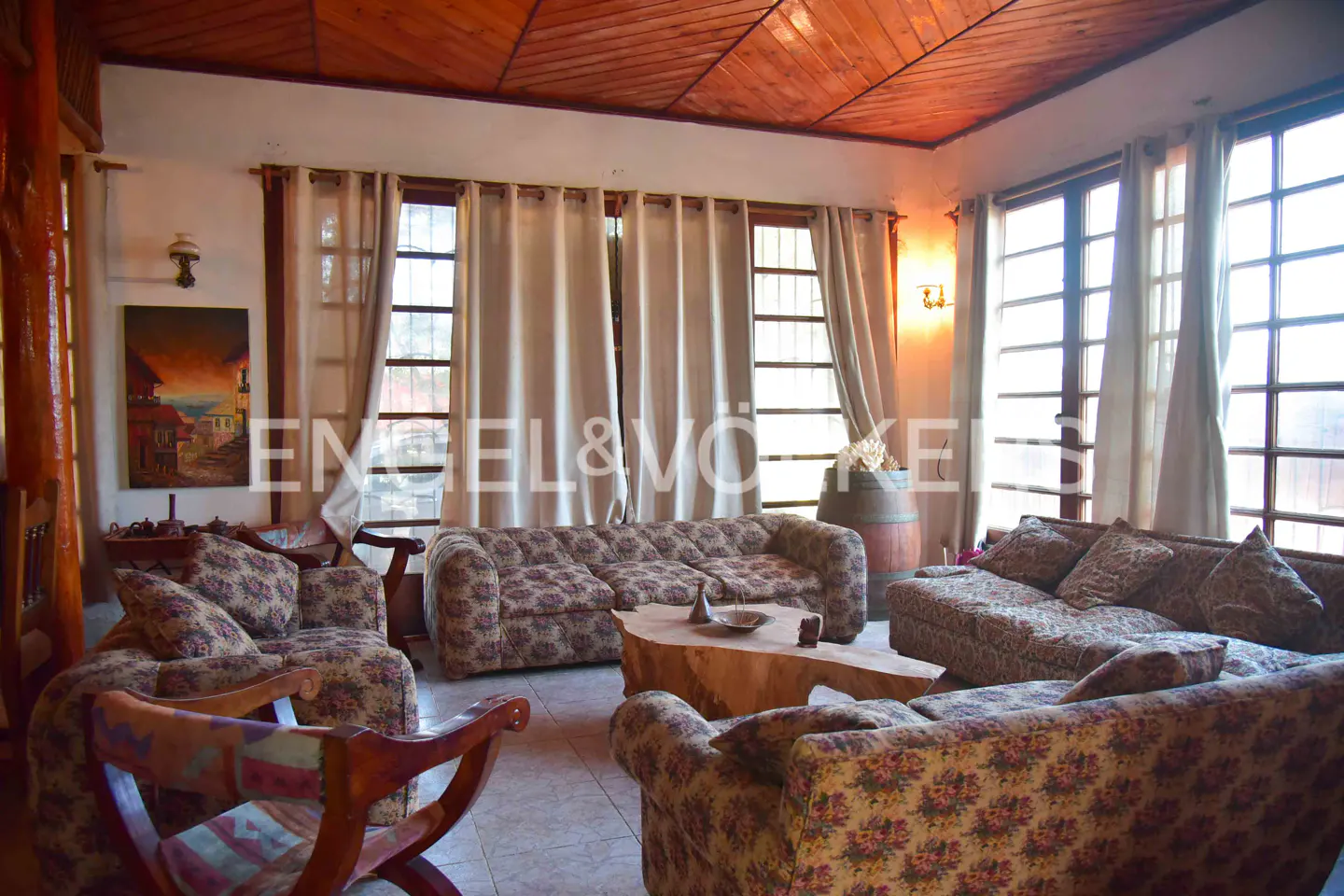 Living room with floral sofas, wood ceiling, and large windows with beige curtains. A rustic wood table sits in the center.