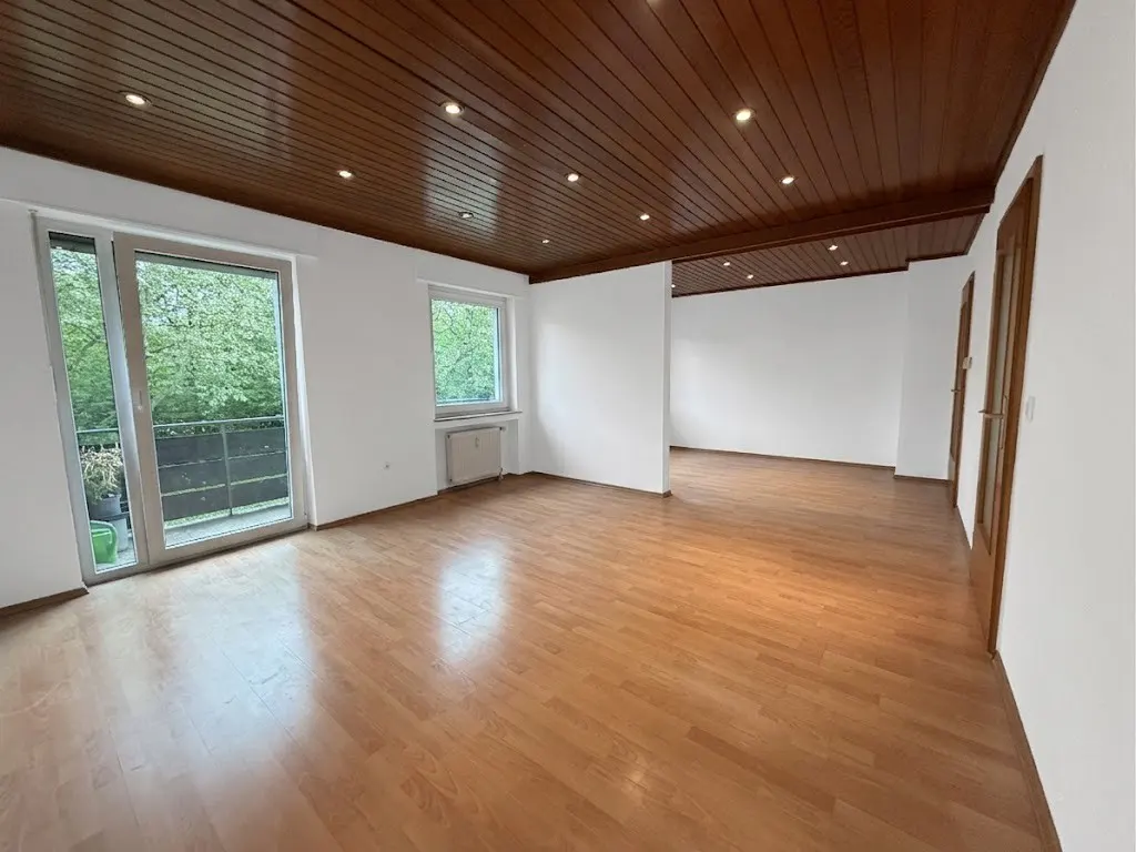 Bright, empty room with wood floors, white walls, and a dark wood paneled ceiling with recessed lighting. A balcony is visible through a sliding glass door.
