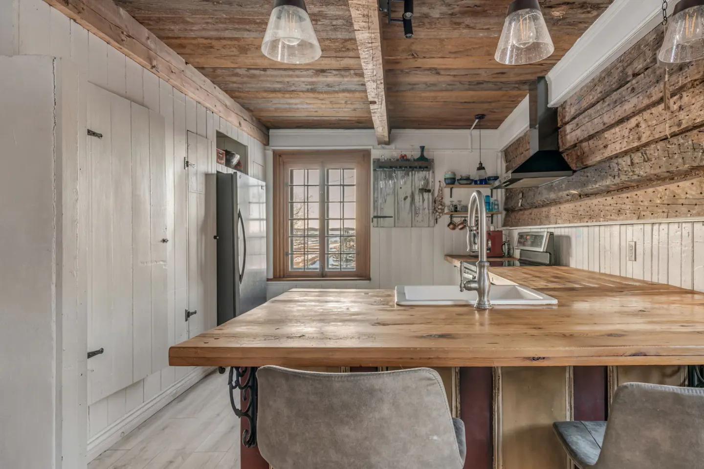 Rustic kitchen with wood ceiling, log walls, and white cabinets. A large wood island with sink and gray chairs is in the foreground.