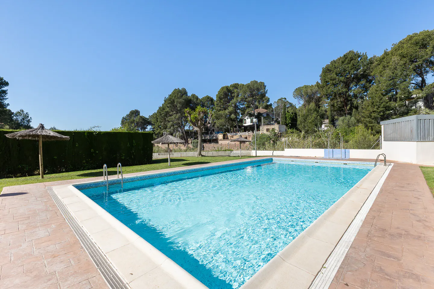 Outdoor pool with clear blue water, surrounded by a brick patio, green grass, and trees under a clear blue sky.