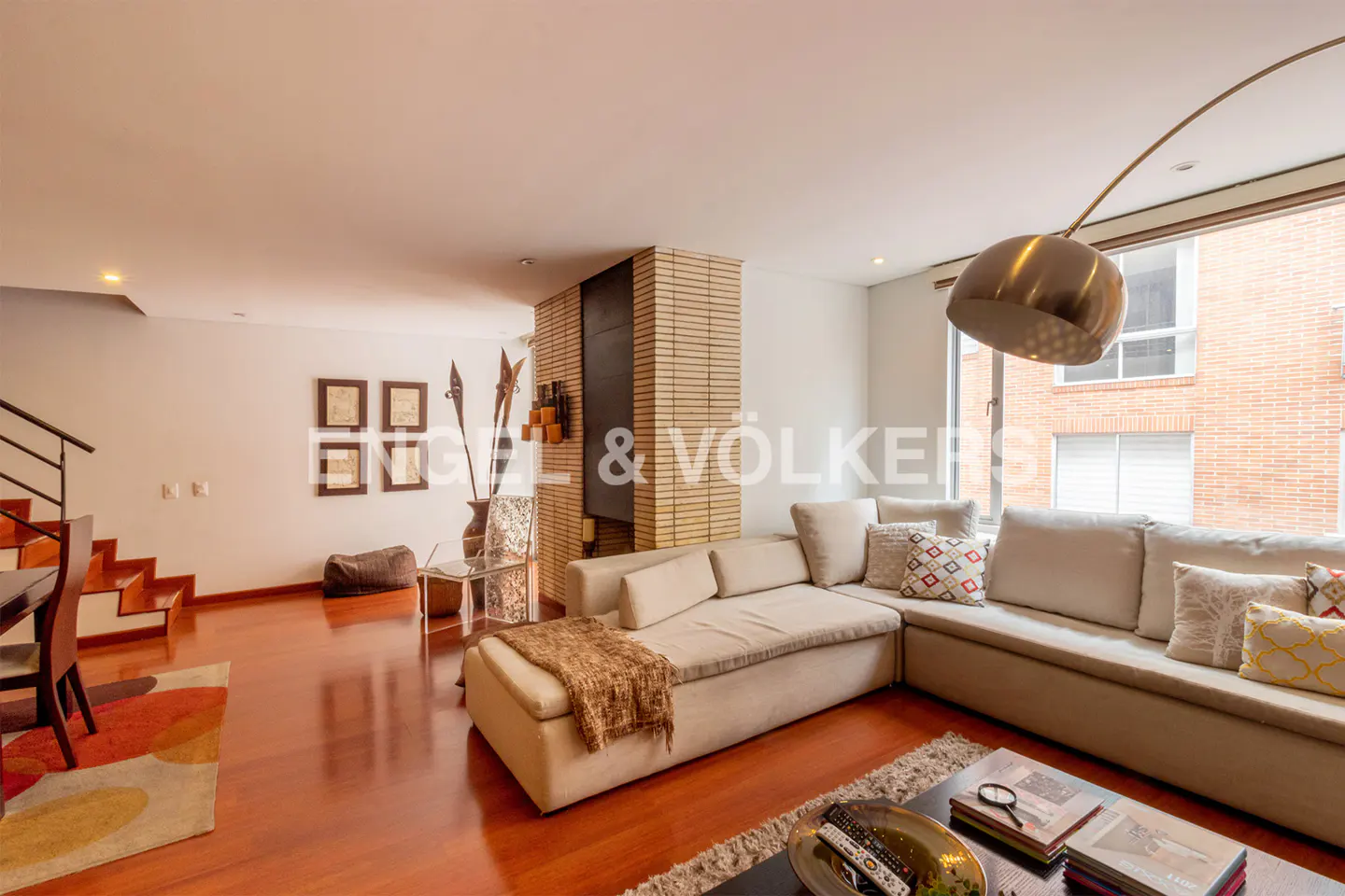 Modern living room with a large beige sectional sofa, hardwood floors, a brick fireplace, and a tall arched floor lamp.