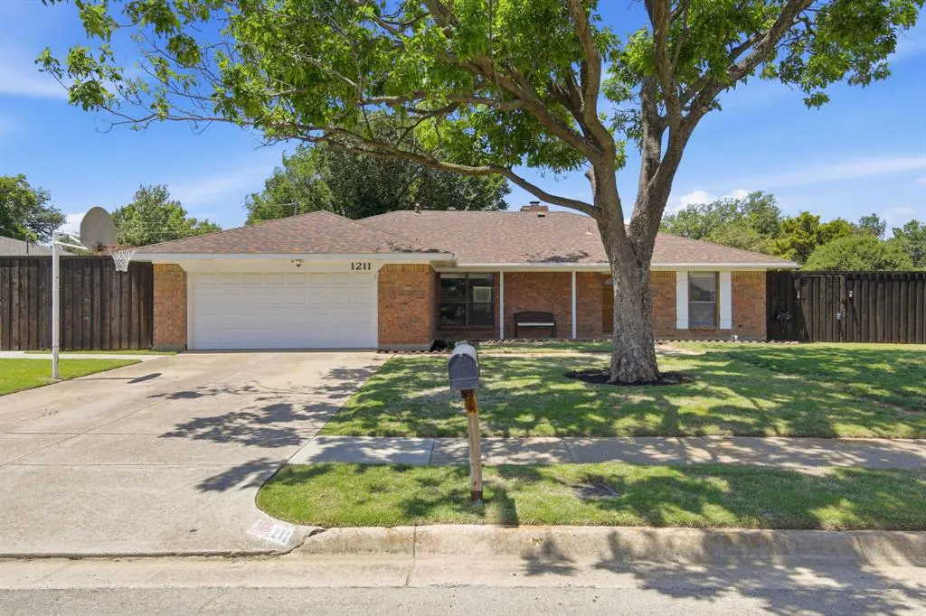 Single-story brick house with white garage door, brown roof, and green lawn under a blue sky. A large tree shades the front.