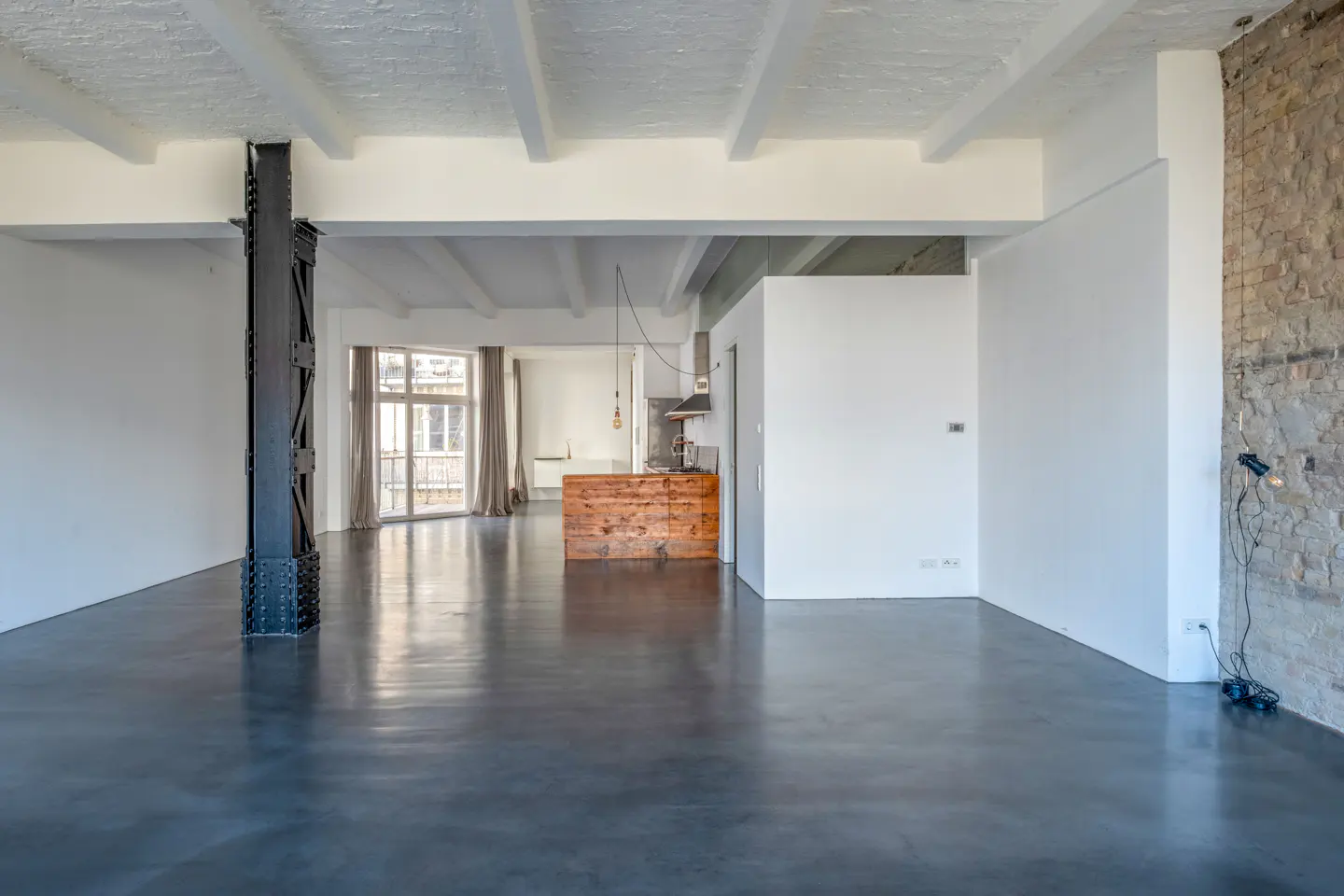 Bright, empty loft with dark floors, white walls, and exposed brick. A black support beam stands near a window with curtains. A wooden kitchen island is visible.