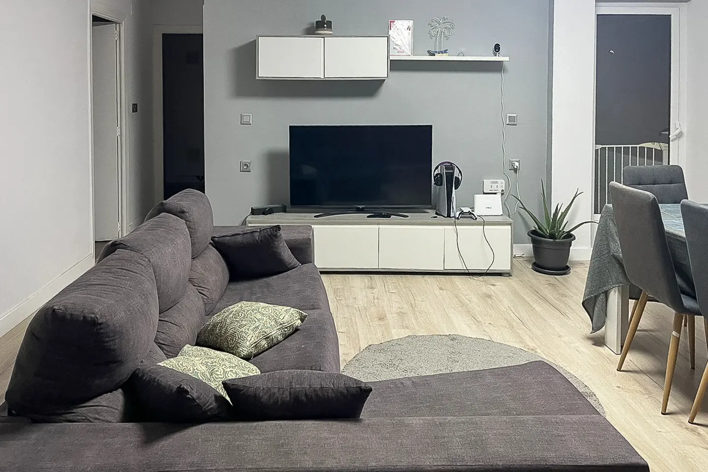 Living room with gray walls, a large gray sectional sofa, a TV on a white media console, and a dining table with gray chairs.