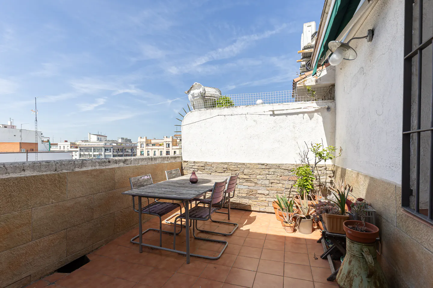 Outdoor patio with a wooden table, chairs, and potted plants. City buildings are visible in the background under a blue sky.