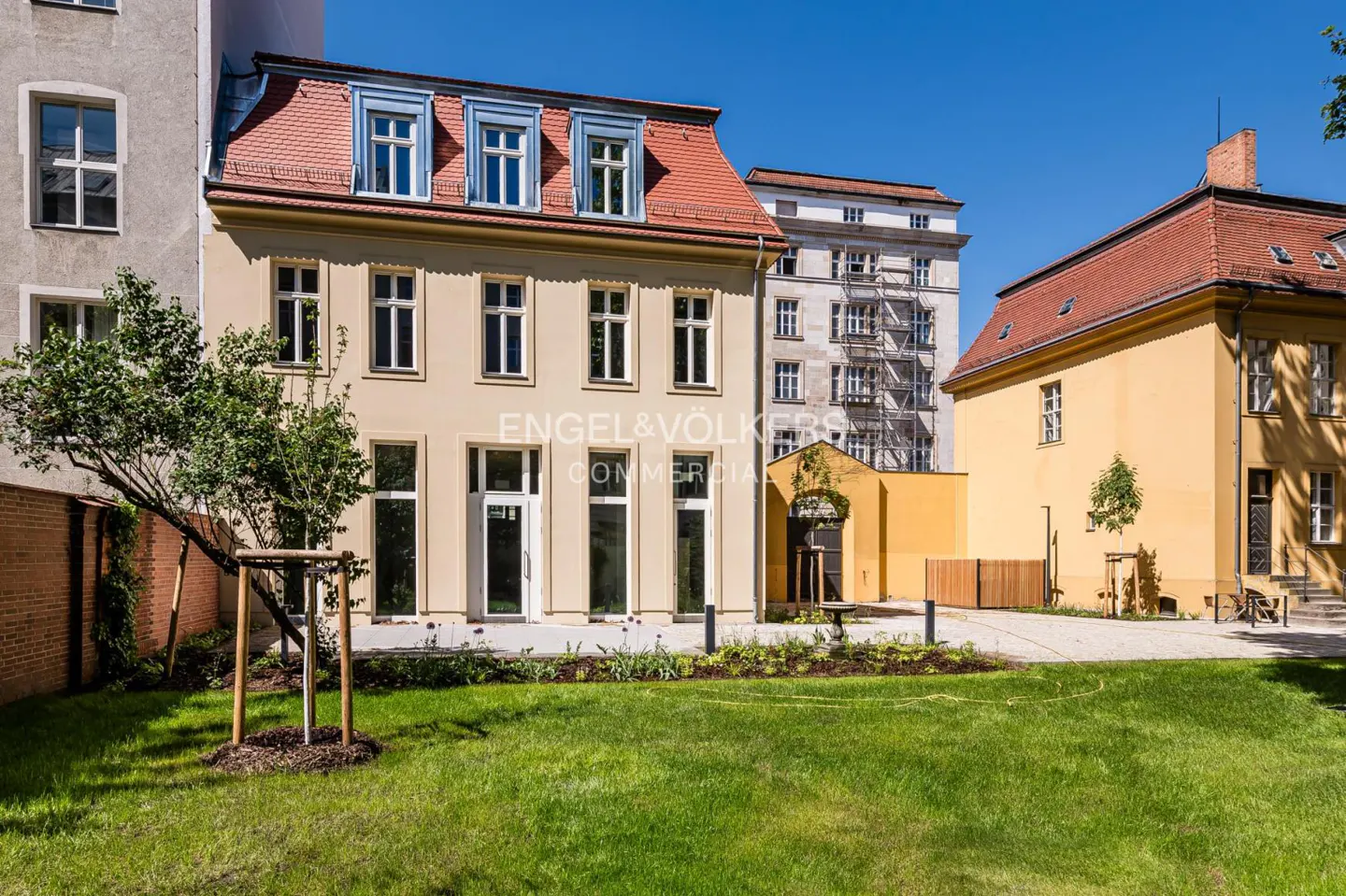 Exterior of a beige building with a red roof and white windows, surrounded by green grass and trees. Engel & Völkers Commercial logo is visible.