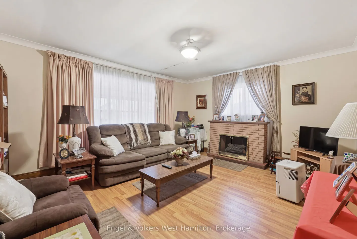 Living room with brown sofa, brick fireplace, wood floors, and beige walls. Curtains frame the windows. A coffee table sits in the center.