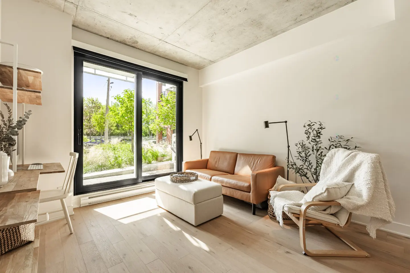 Bright living room with a brown leather sofa, white ottoman, and a wooden chair with a white blanket. A desk sits by a large window.