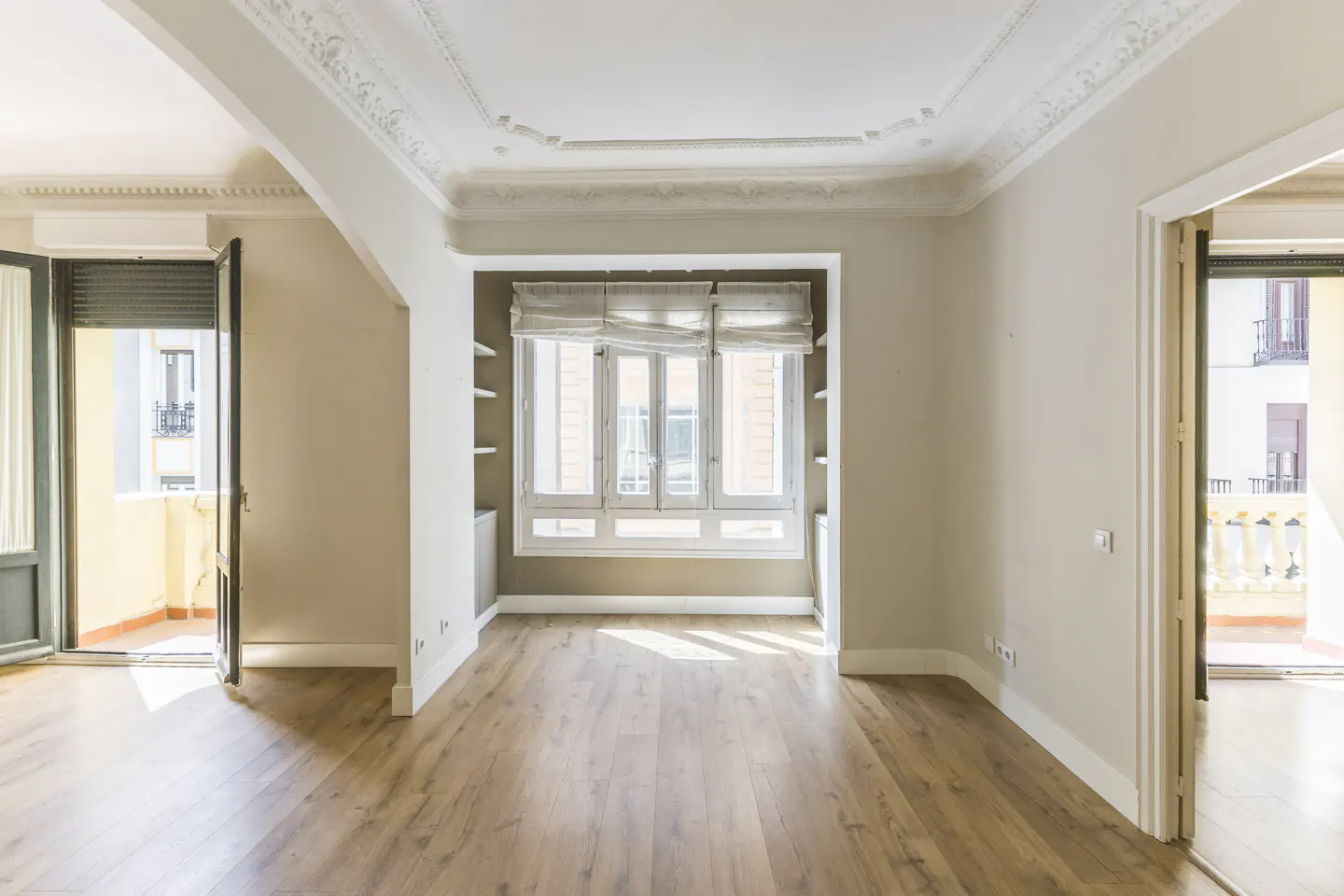 Empty room with wood floors, beige walls, and white trim. A window with blinds and two open doors lead to balconies.