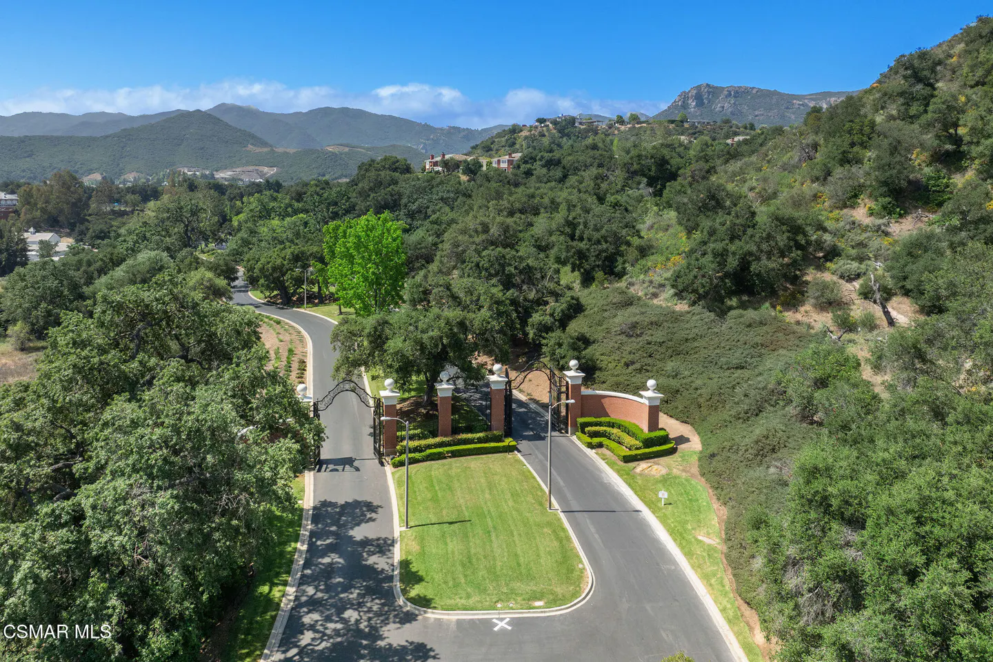 Gated entrance to a property with a long driveway, green trees, and mountains in the background.