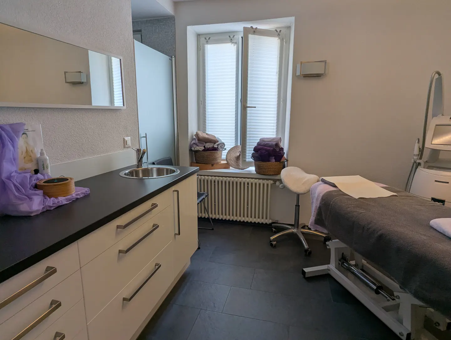 Spa room with a massage table, white cabinets, sink, and window with baskets of towels.
