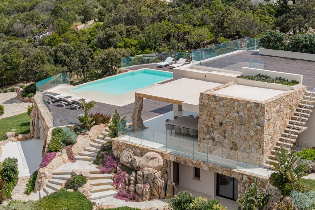 Aerial view of a modern stone villa with a pool, deck, outdoor dining area, and landscaped stairs set in a wooded area.
