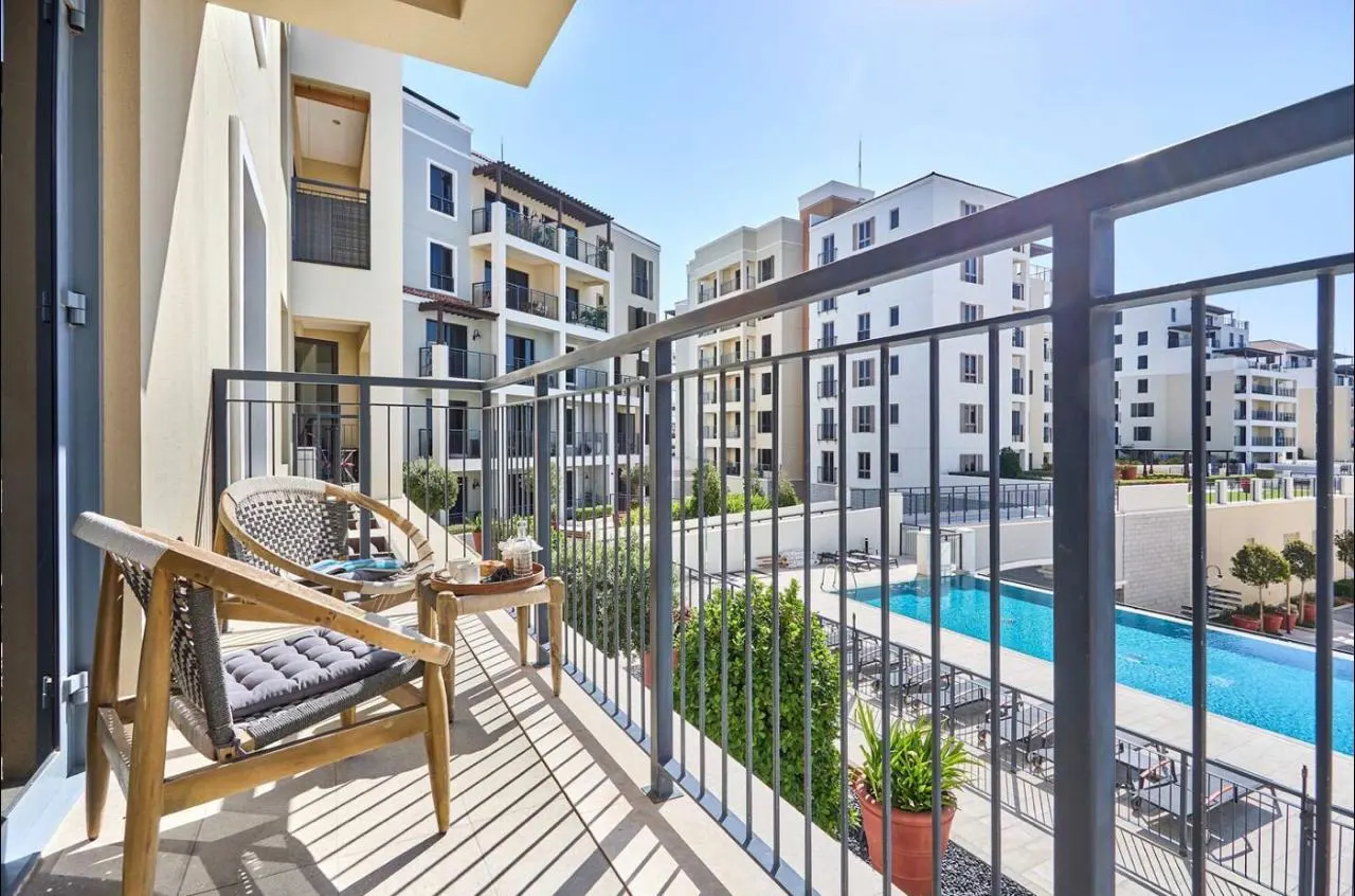 Balcony view with two chairs, a small table, and a metal railing overlooking a pool and apartment buildings on a sunny day.