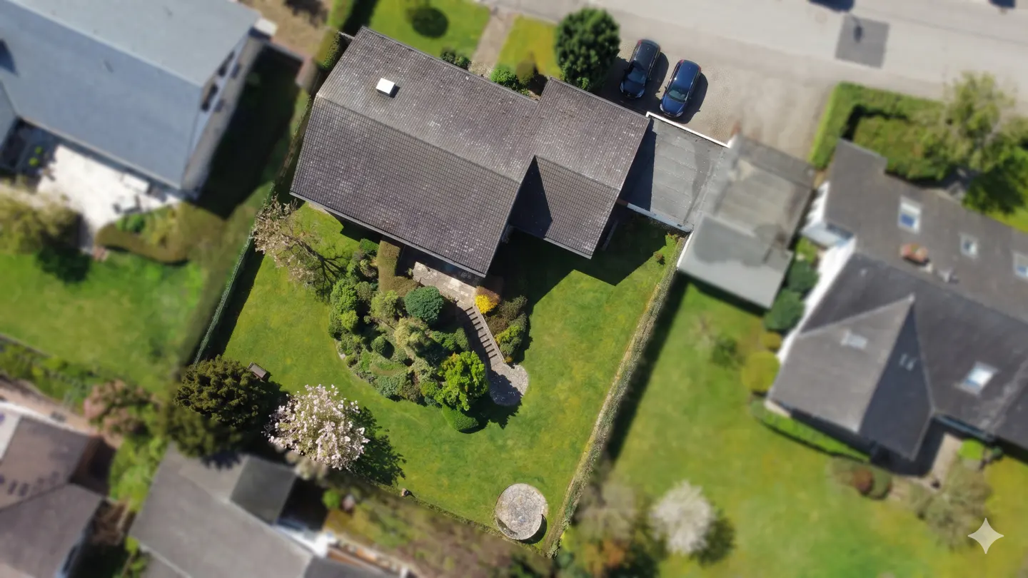 Aerial view of a single-story house with a gray roof, green lawn, and landscaped garden. Two cars are parked nearby.