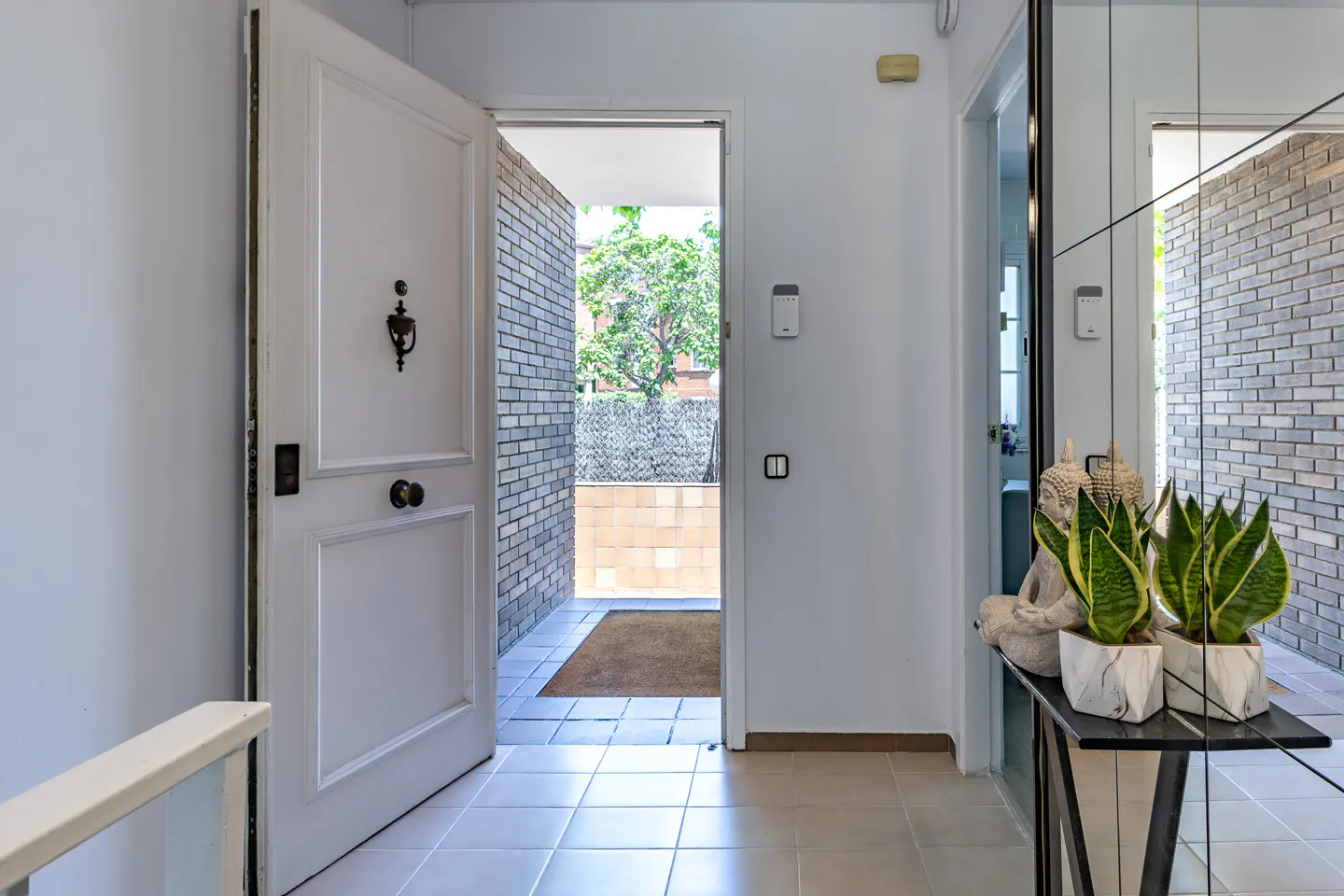 Open white front door leading to a brick-walled patio, with a Buddha statue and snake plants on a console table inside.