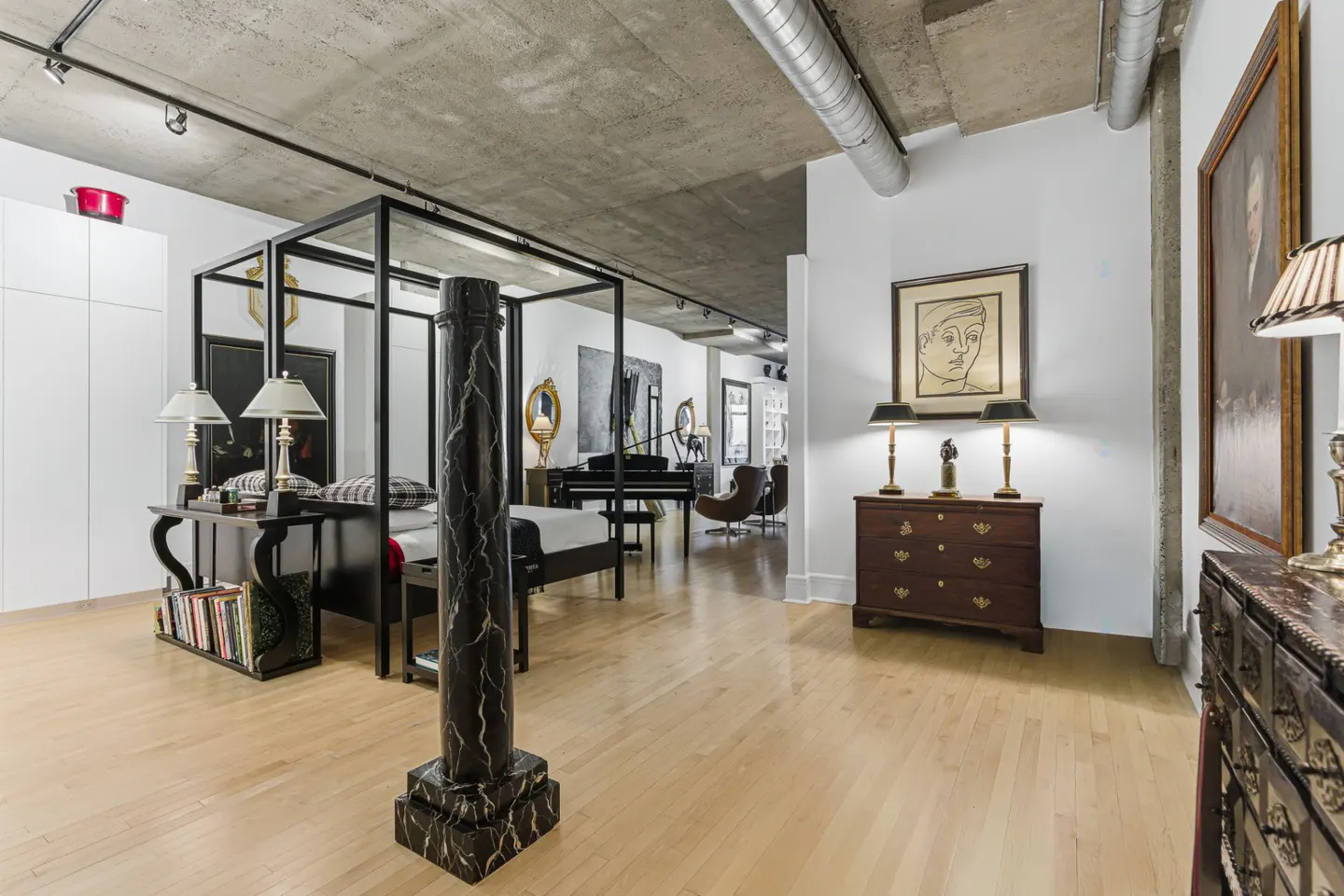 A modern apartment interior with a four-poster bed, piano, art, and wood floors. The ceiling is concrete with exposed pipes.