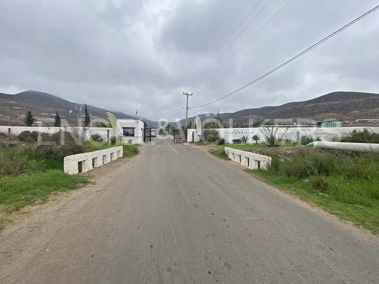 Entrance to La Varilla, a gated community. A gray road leads to a white wall with the community's name. Mountains are in the background.