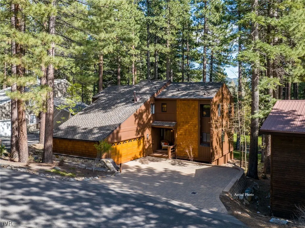 A two-story brown house with a gray roof is surrounded by tall green trees. A paved driveway leads to the garage.
