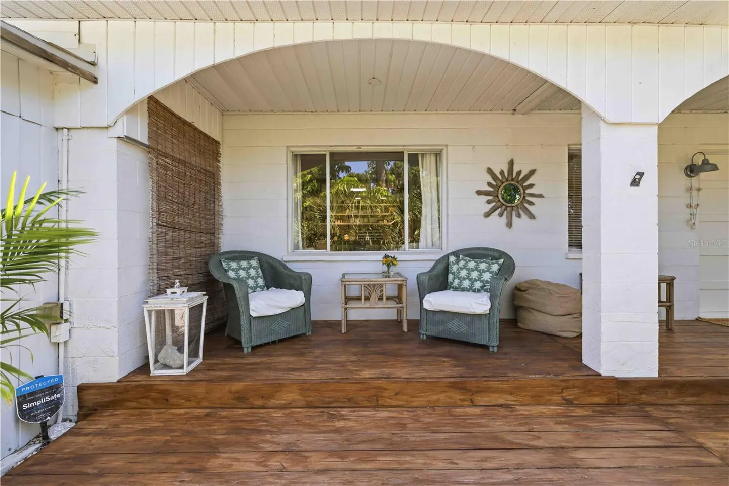 Covered porch with two green wicker chairs, white cushions, and patterned pillows. A small table sits between them, with a window and sunburst mirror behind.