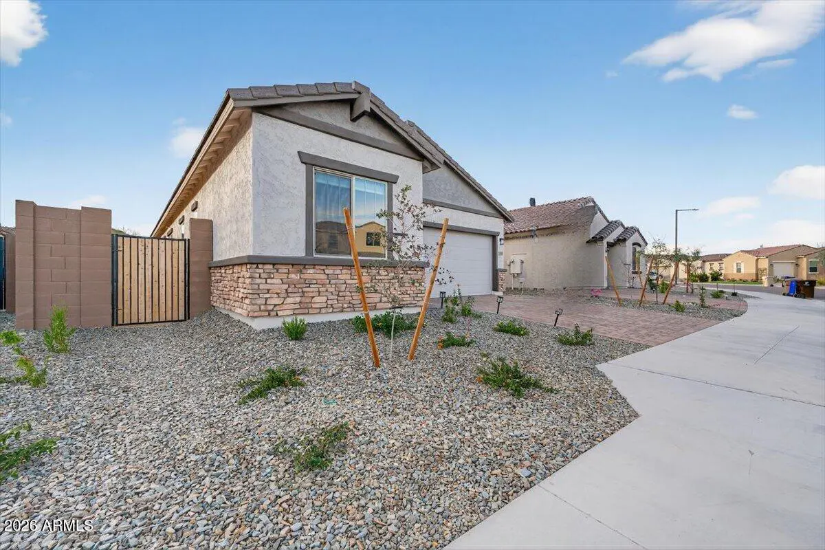 A beige single-story house with a stone facade and a gray roof, set in a gravel yard with a wooden gate.