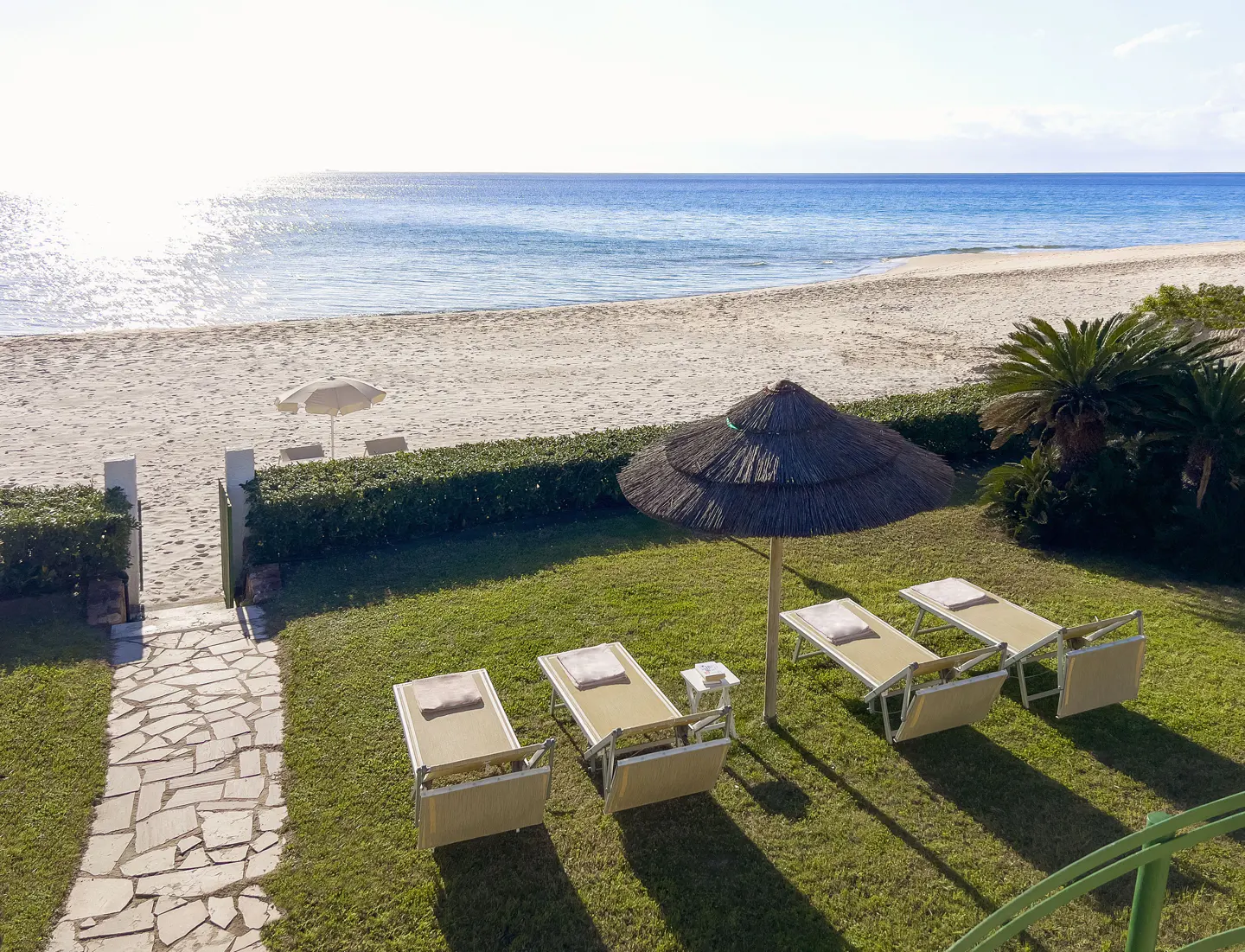 Beachfront property with lounge chairs on green grass under a thatched umbrella, leading to a sandy beach and blue ocean.