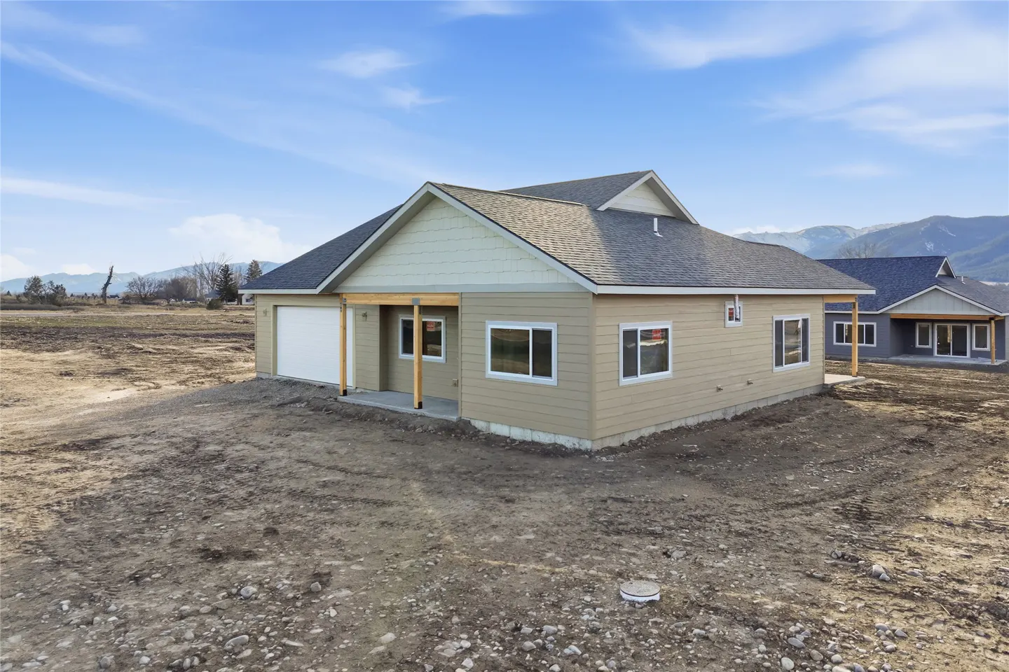 A tan single-story house with a gray roof and white garage door sits on a dirt lot under a blue sky.