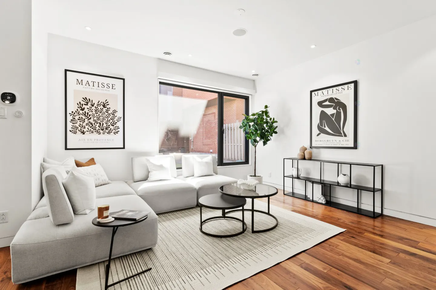 Bright living room with white walls, wood floors, and a gray sectional sofa. Two Matisse prints hang on the walls. A black shelving unit and round nesting tables add modern touches.