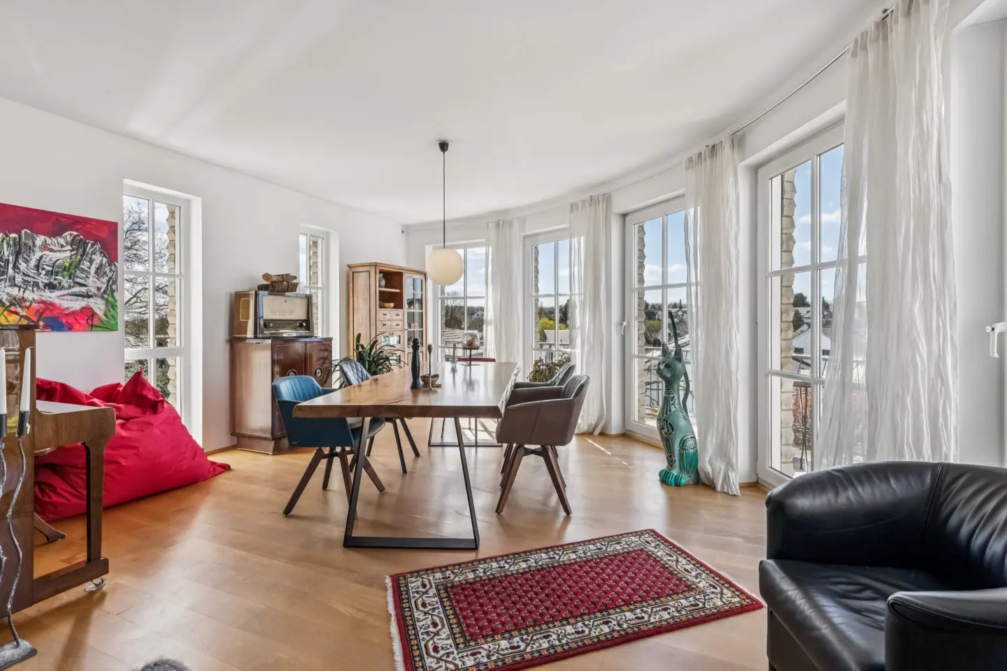 Bright dining room with wood floors, a long wood table with chairs, and large windows with white curtains. A red rug and beanbag add color.