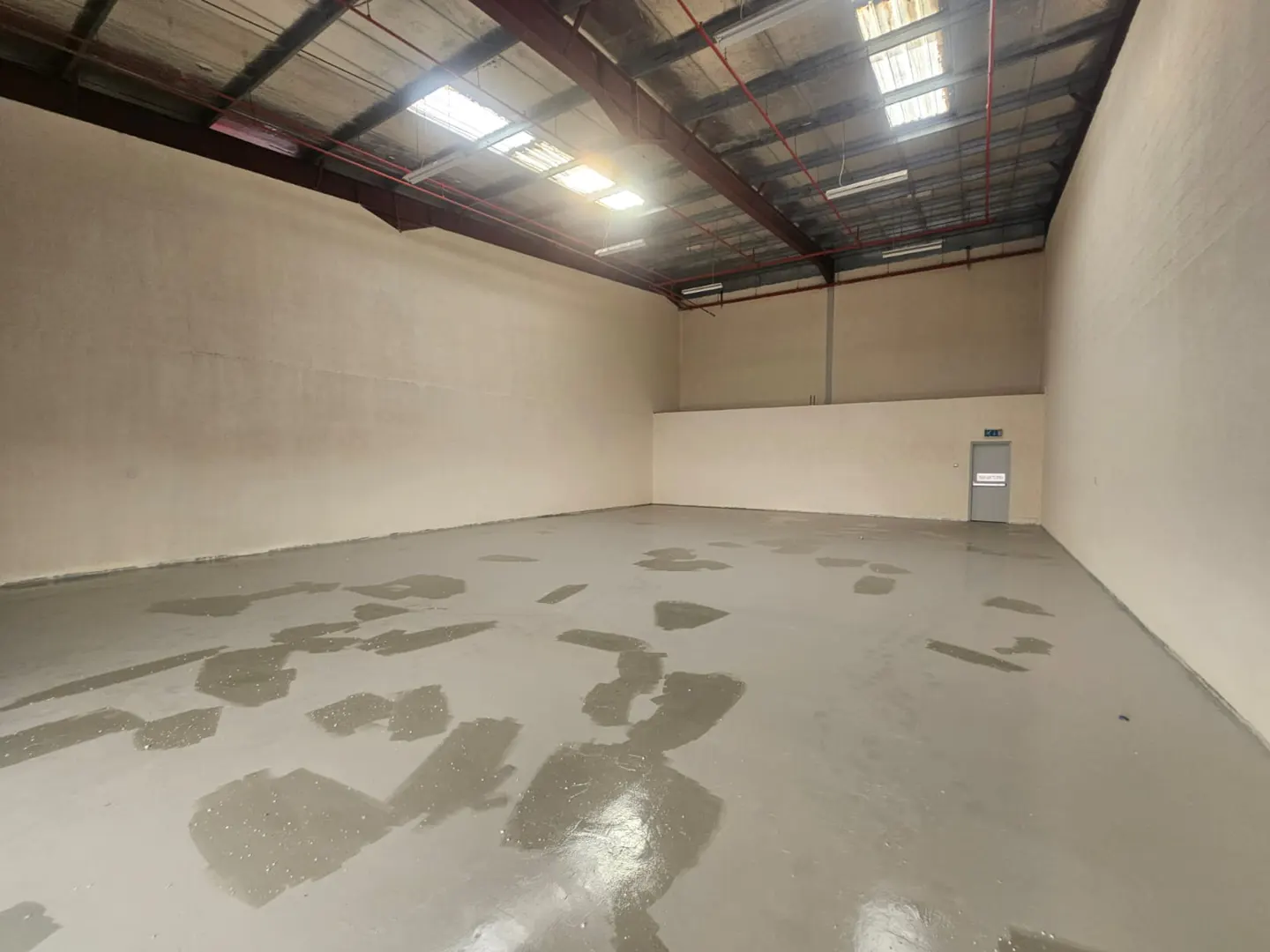 Empty warehouse interior with gray floor, beige walls, and metal ceiling beams. A door is visible in the back right corner.