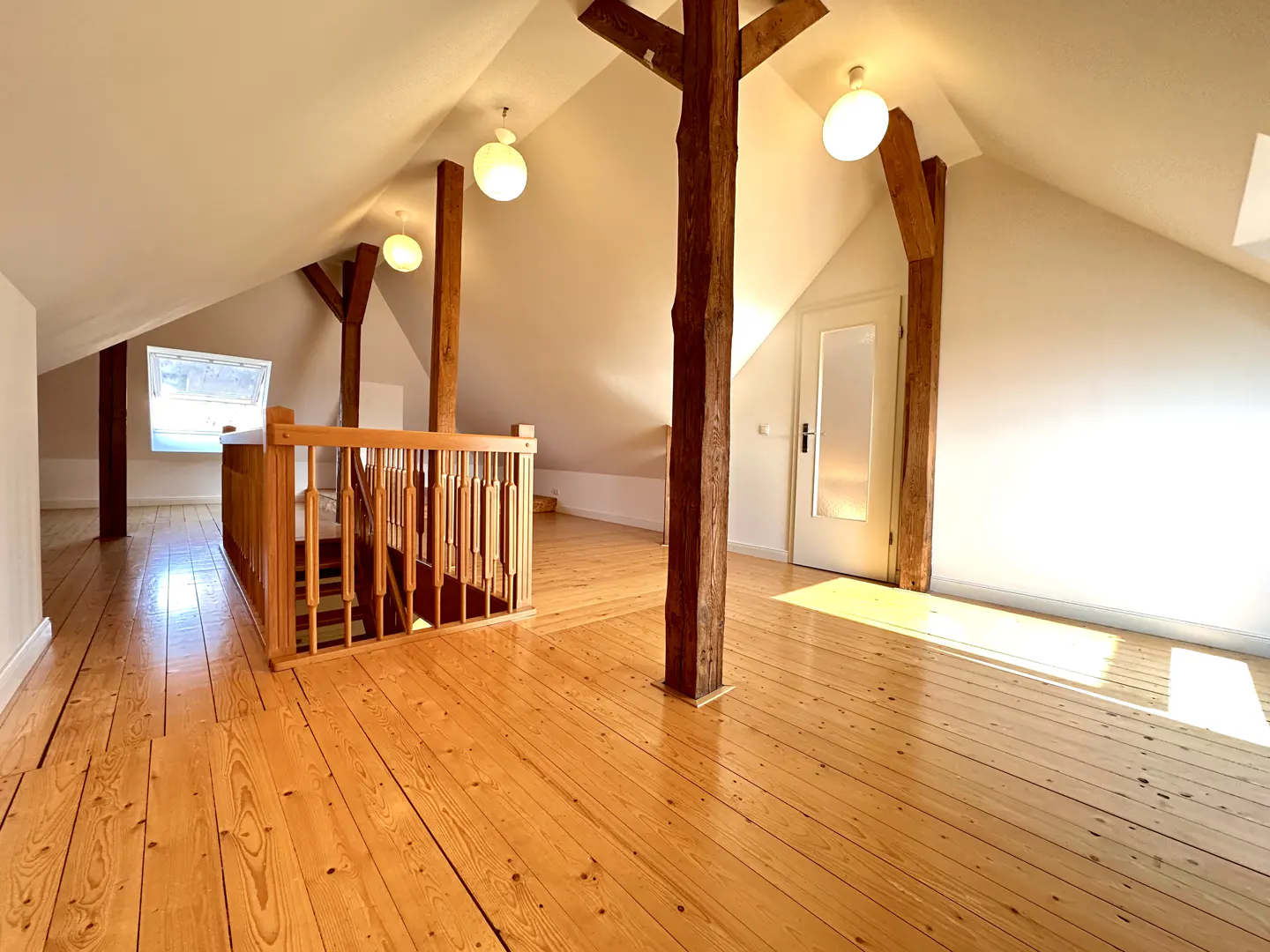 Attic room with wooden floors, white walls, and exposed wooden beams. A wooden railing overlooks a lower level. A white door is visible.