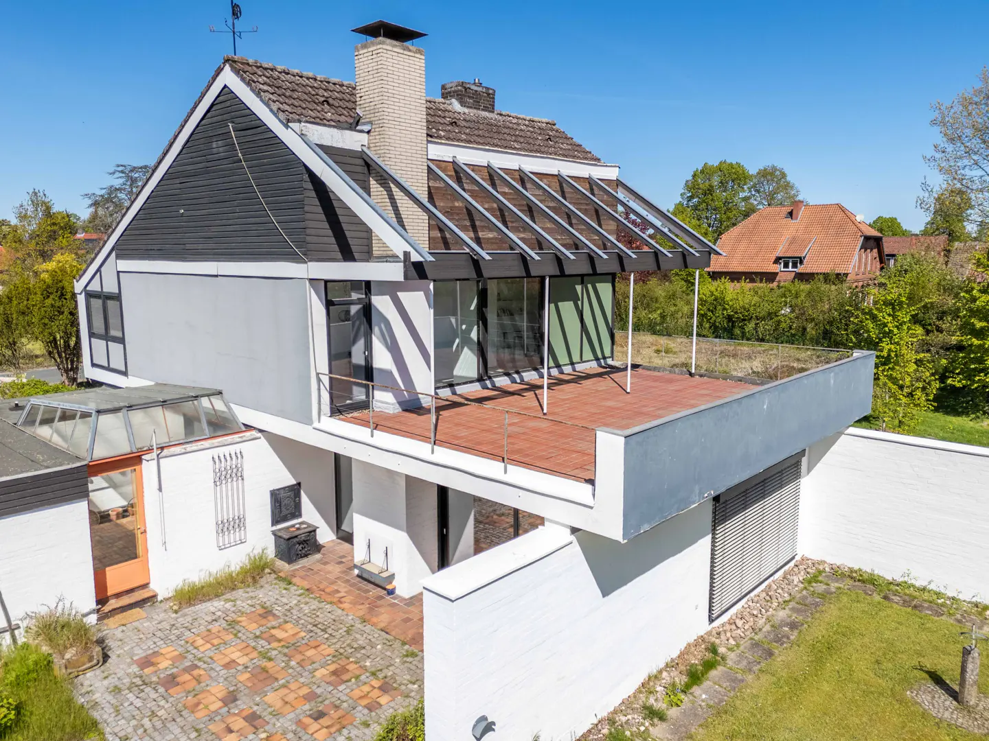 Aerial view of a modern two-story house with a gray roof, white walls, and a brick patio. A glass-covered terrace is on the second floor.