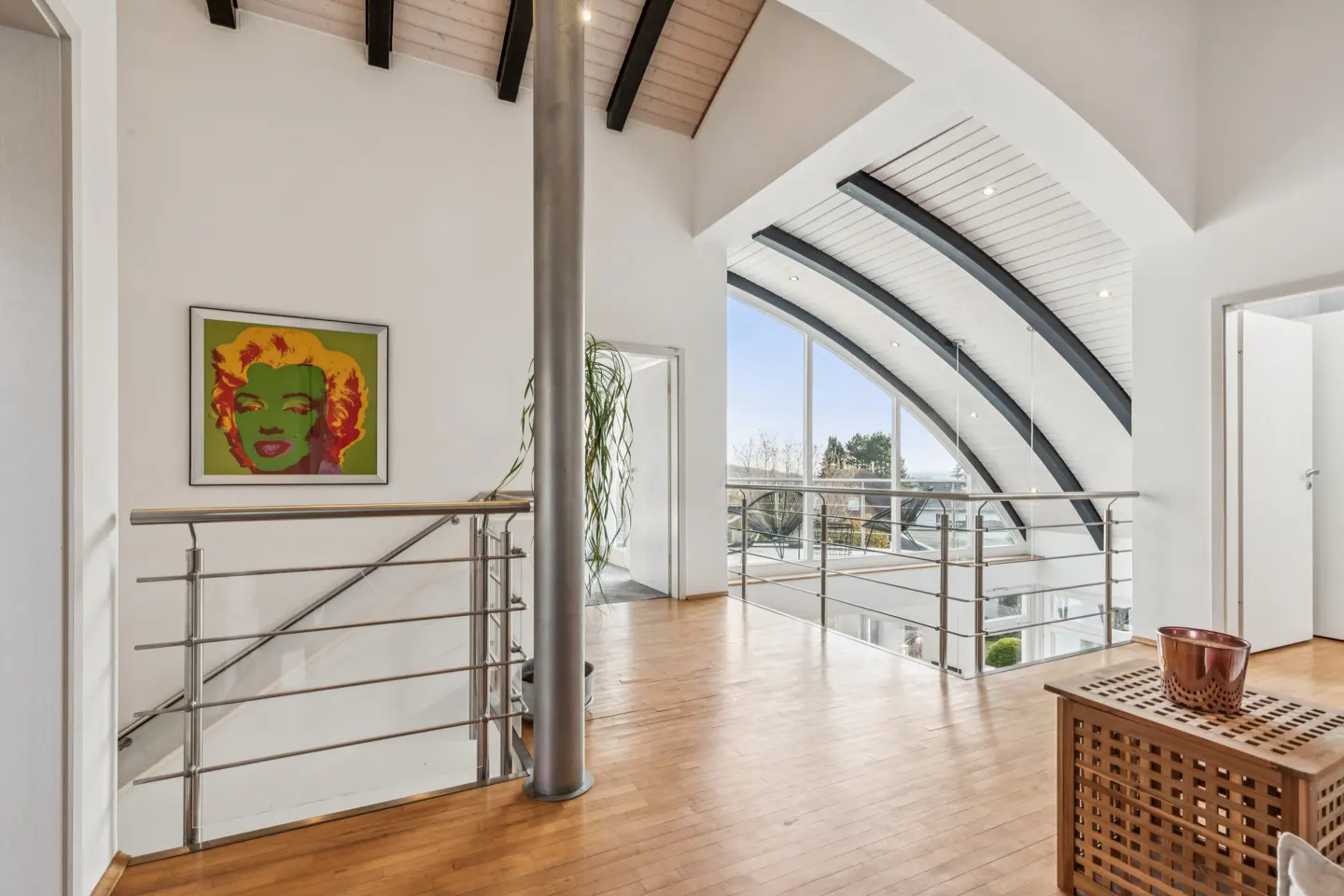 Bright, modern home interior with wood floors, white walls, and a vaulted ceiling with black beams. A colorful portrait hangs near a metal railing.