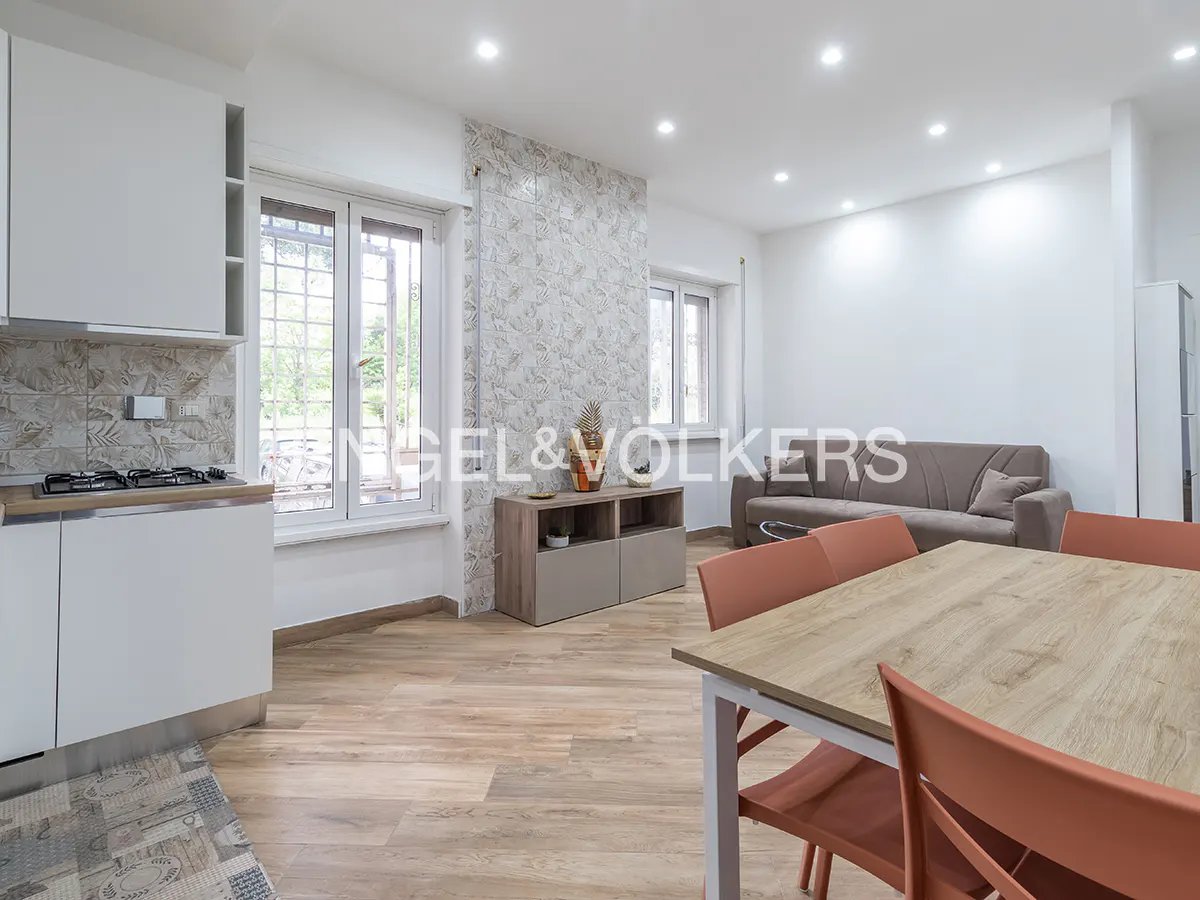 Open-concept living space with wood floors, white kitchen cabinets, dining table with orange chairs, and a gray sofa.