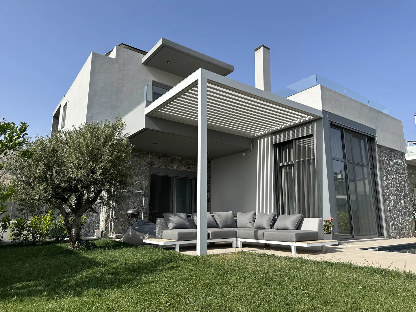 Modern two-story house with a gray pergola over a patio with a gray sofa and green lawn.