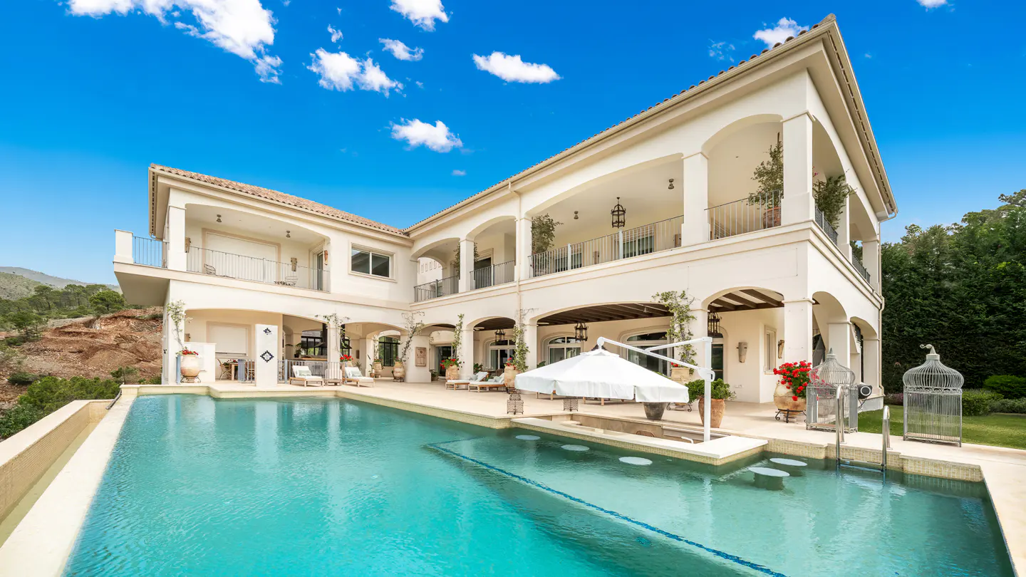 Luxury home with a pool. The white stucco building has arched balconies and a red tile roof. Blue sky above.