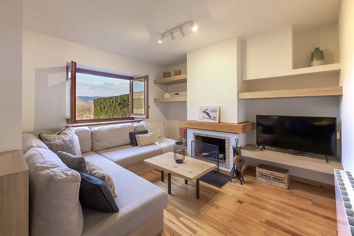 Living room with a beige sectional sofa, wood floors, and a fireplace. A window shows a view of trees and hills.