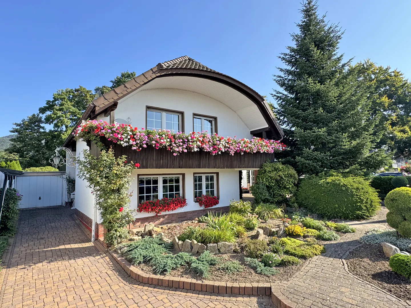 Two-story white house with brown trim and a curved roof. Balcony and window boxes are overflowing with pink and red flowers. Brick driveway and lush landscaping.