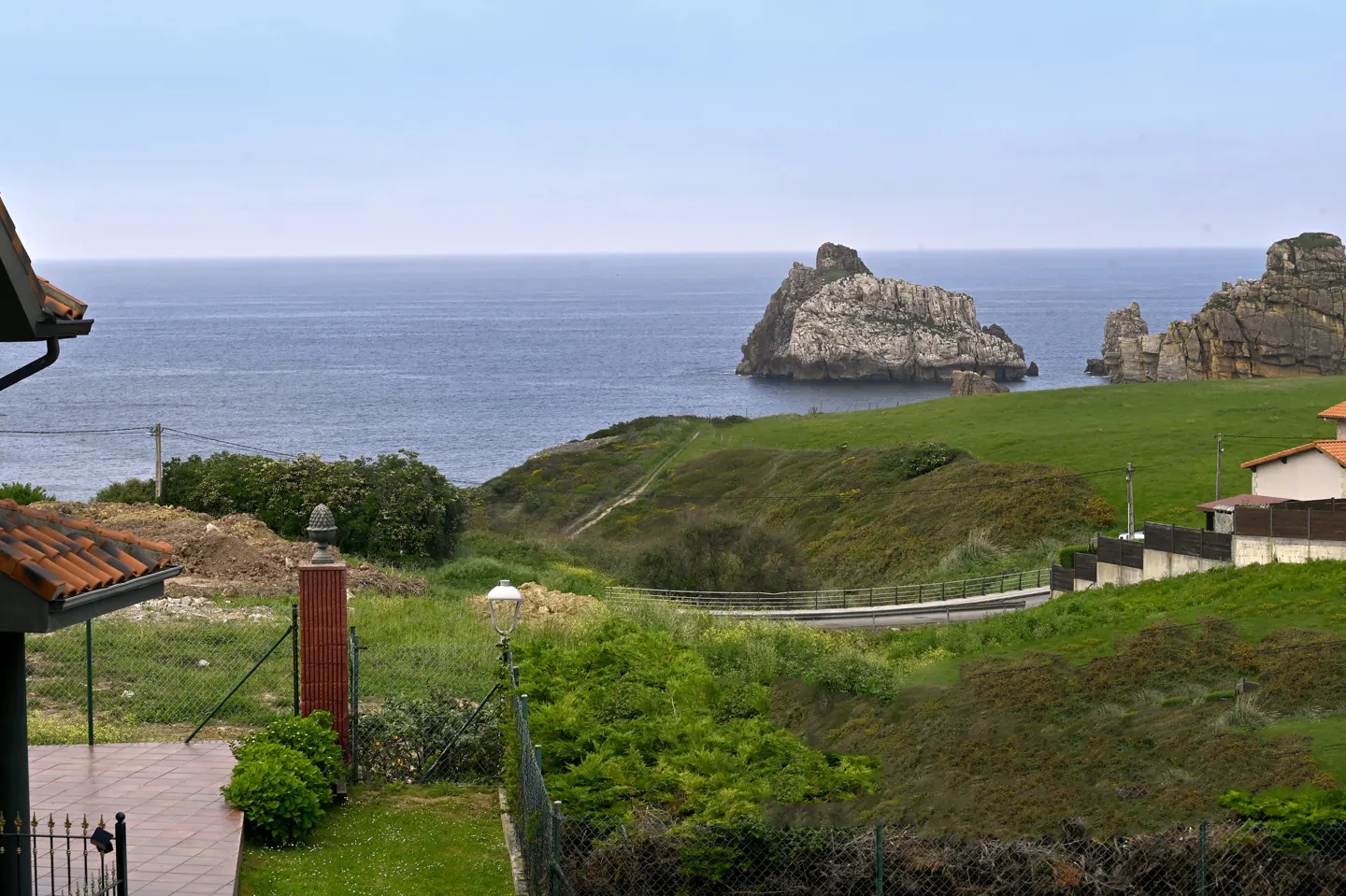 Ocean view from a property with green hills, rock formations in the water, and a red-tiled roof visible.