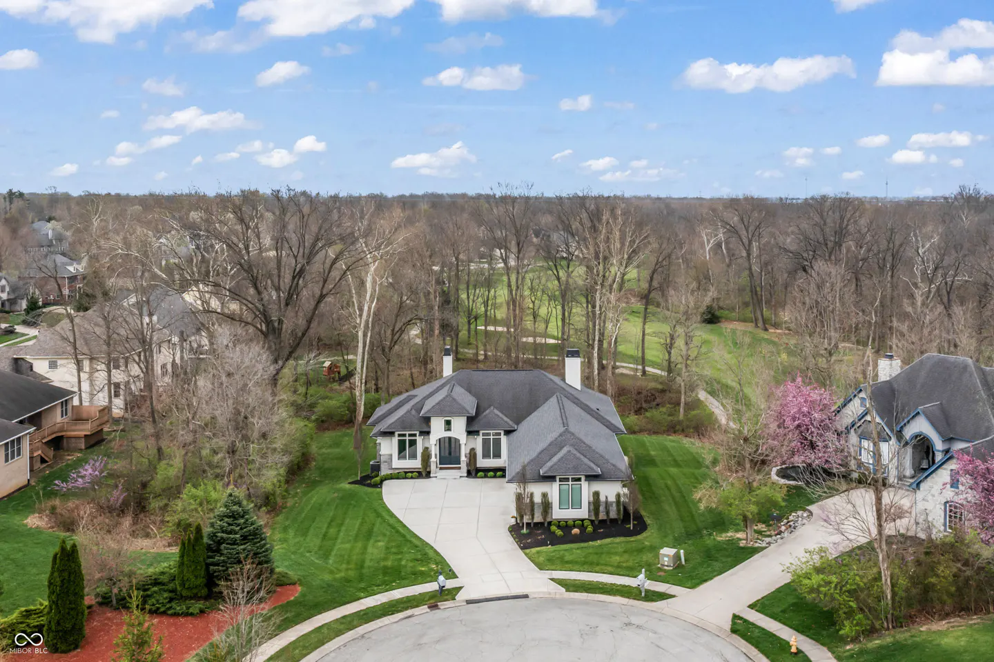 Aerial view of a white, single-story house with a gray roof, surrounded by green lawns and trees under a blue sky with clouds.