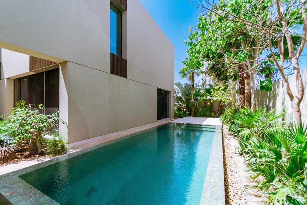 A modern home with a turquoise pool, surrounded by lush greenery and a clear blue sky.