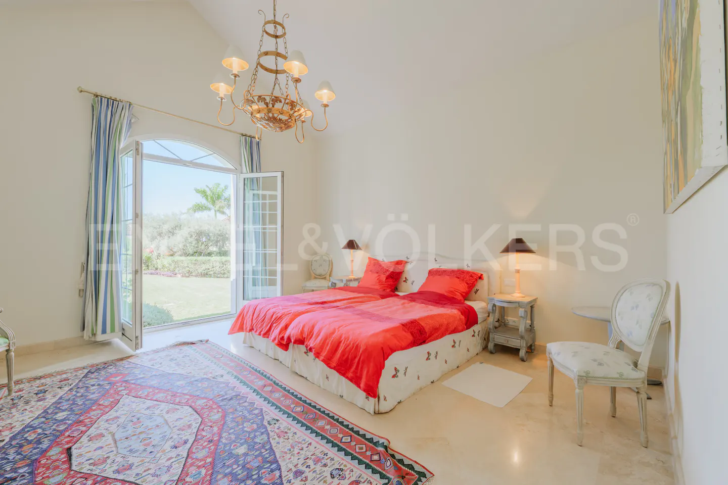 Bedroom with two twin beds, red bedding, chandelier, and open doors to a green lawn.